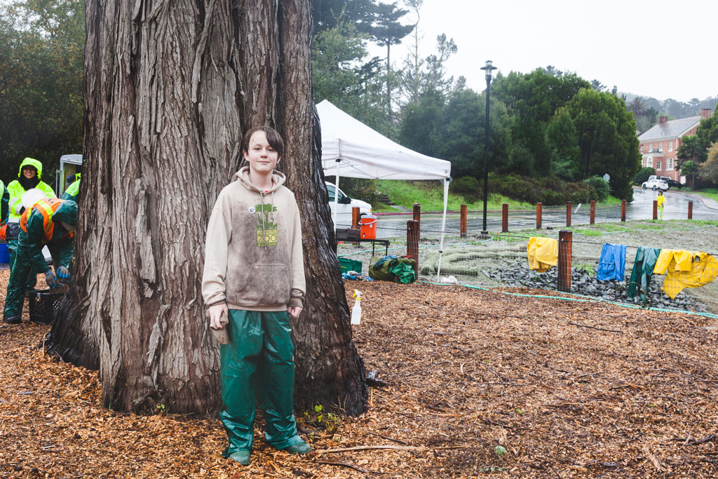 Young man in San Francisco Zoo sweatshirt and rain pants in front of tree