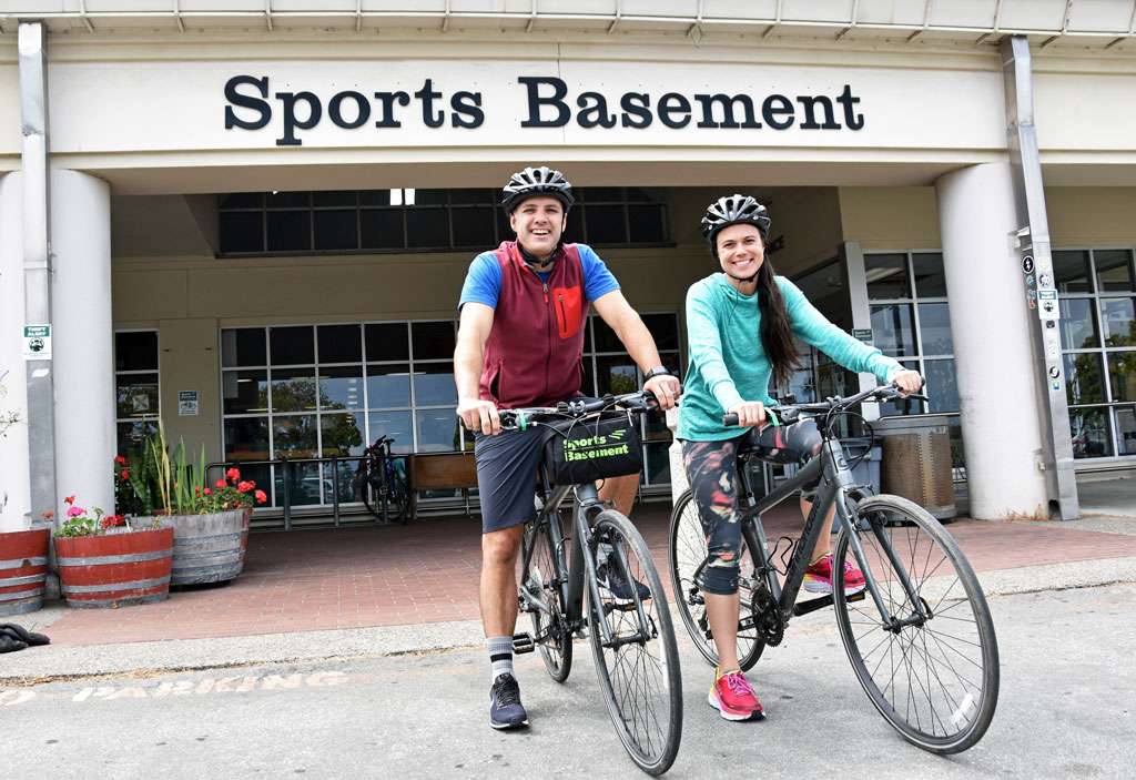 Man and woman in helmets on rented bikes in front of Sports Basement.
