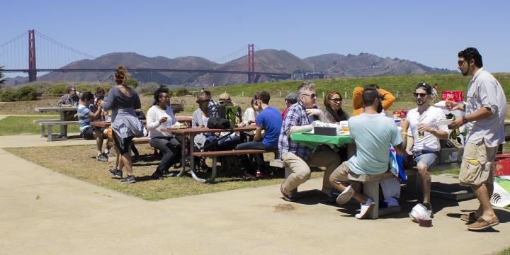 PIcnickers on picnic tables at Crissy Field with Golden Gate Bridge in background.