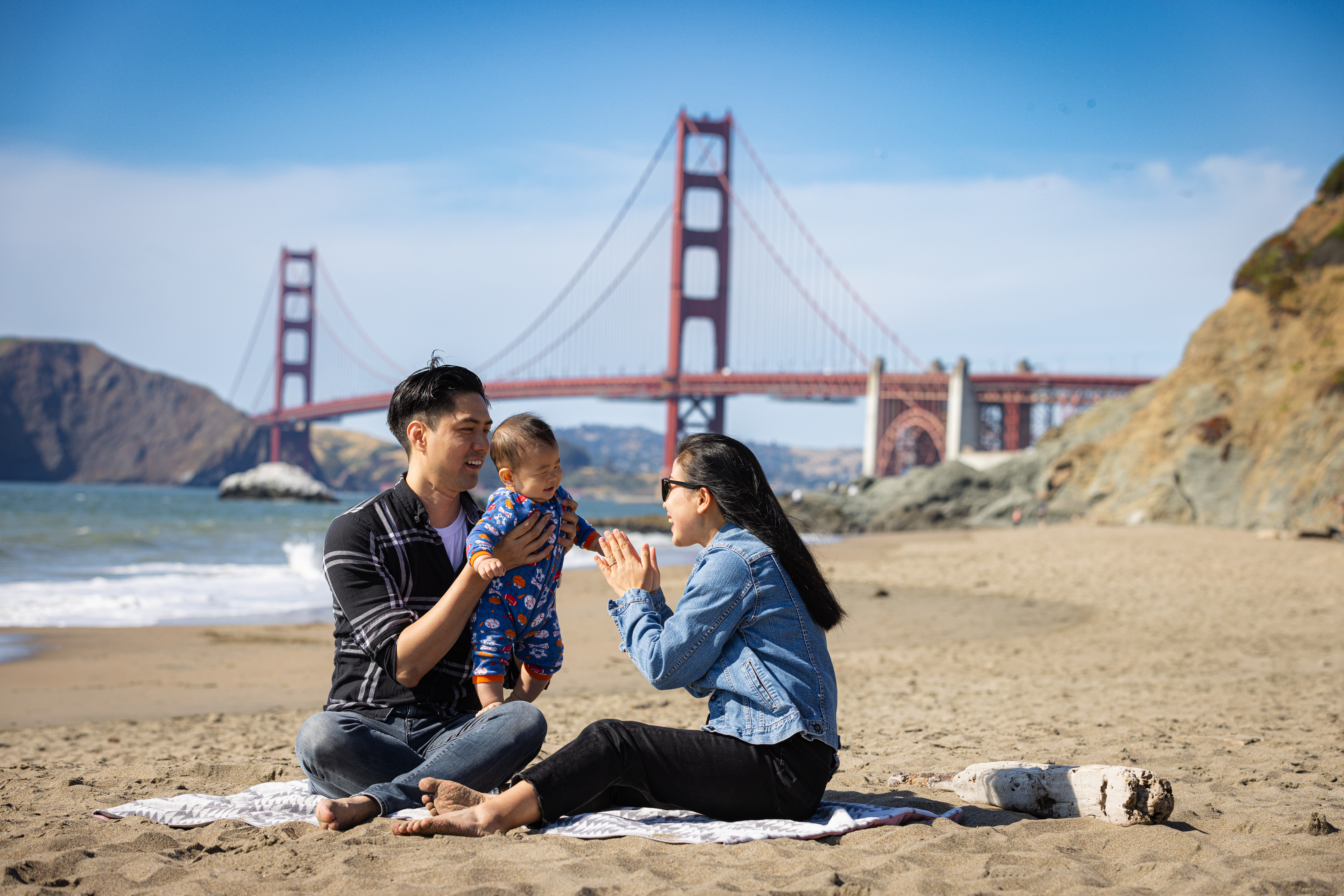 Young couple playing on a blanket with their baby at Baker Beach with the Golden Gate Bridge in the background