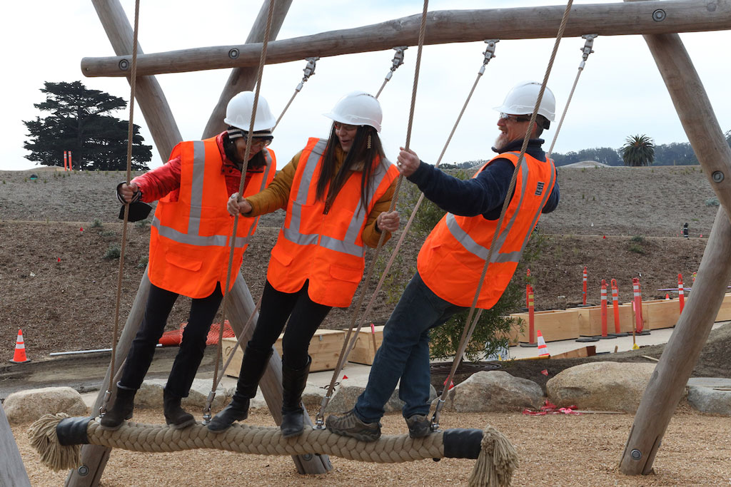 Three people in safety vests and helmets standing and holding on rope swings