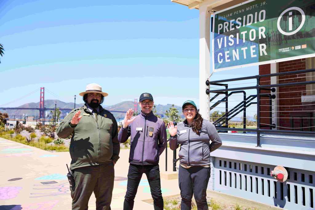 Three people, one with Park Ranger, one wearing Parks Conservancy clothing, and one person with Presidio Trust gear stand outside the Presidio Visitor Center and wave at camera.