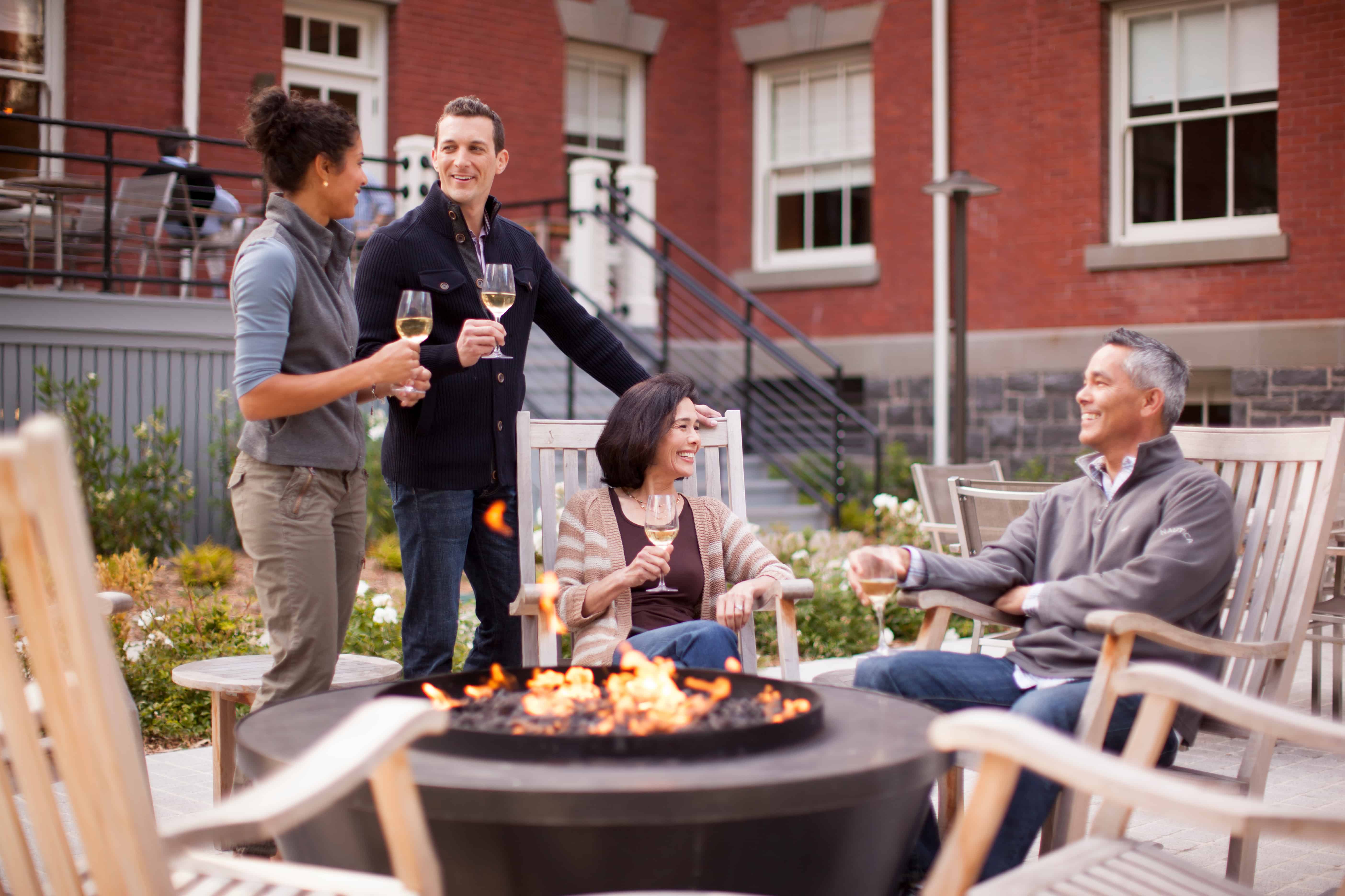 group of 4 people standing and sitting around a fire pit with glasses of wine.