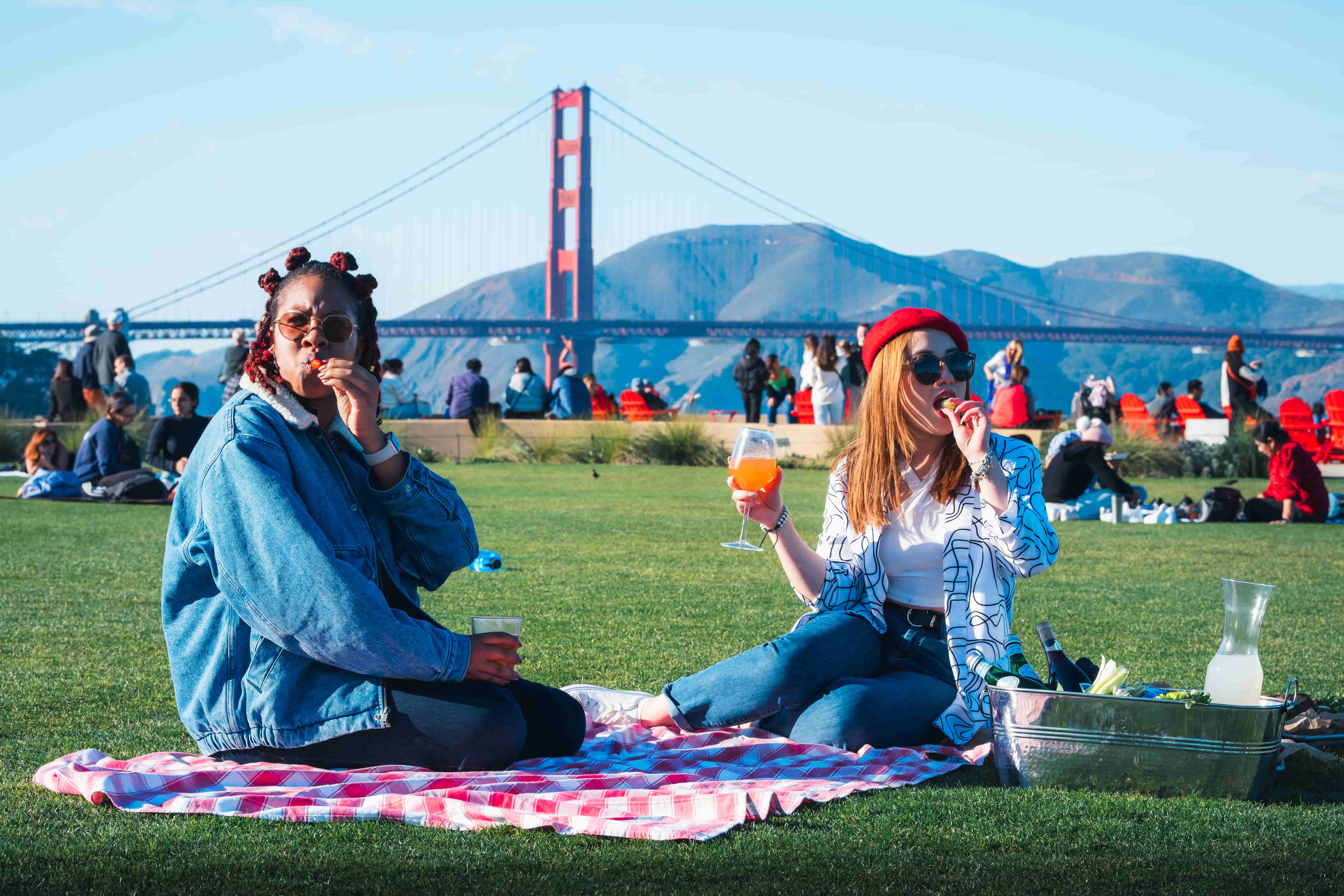 Two women having a picnic on a checkered blanket at the Presidio Tunnel Tops with the Golden Gate Bridge in the background.