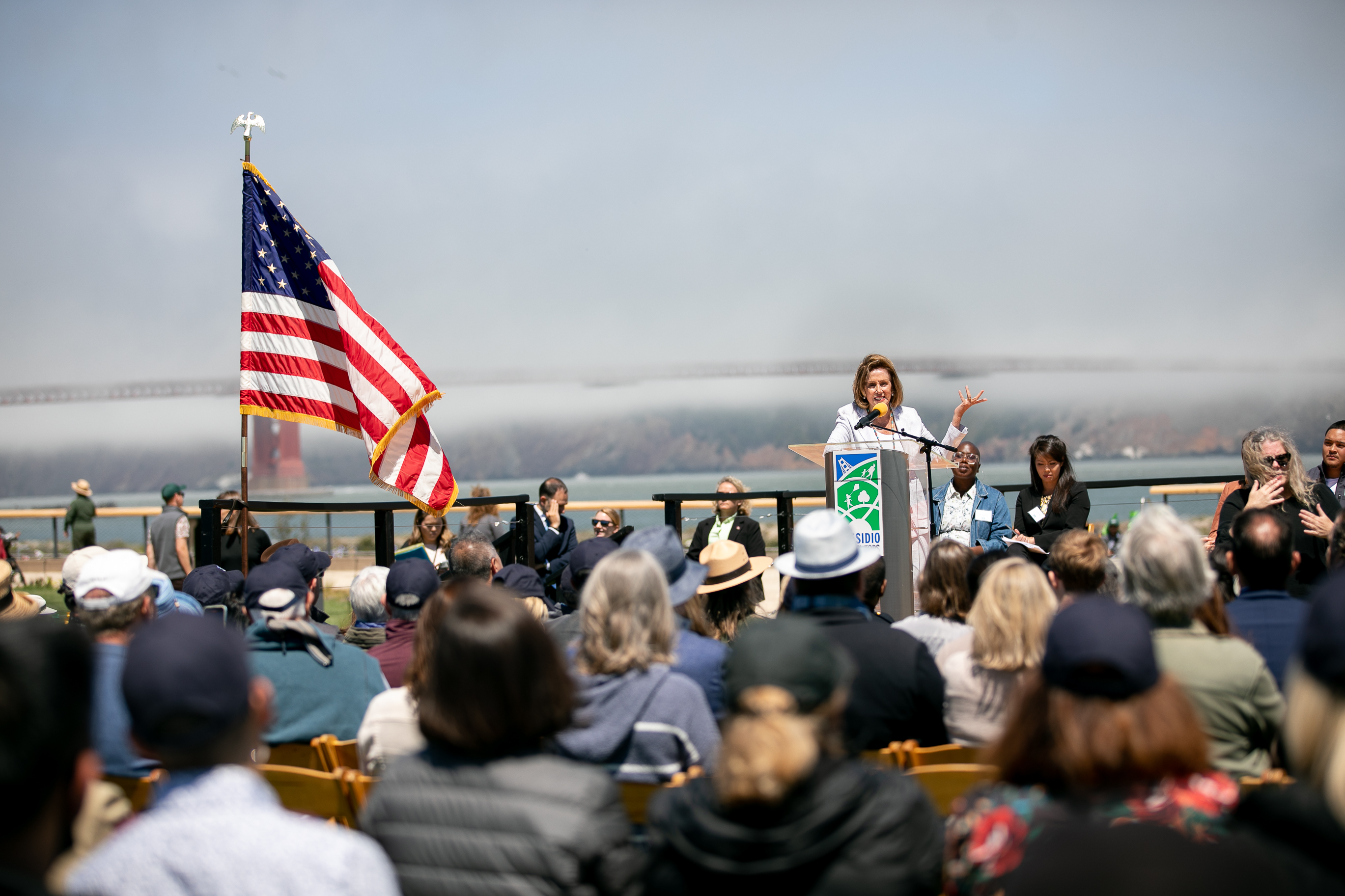 Image of Nancy Pelosi speaking at a podium with a crowd of people listening in front of her and the Golden Gate Bridge hidden in fog behind her.
