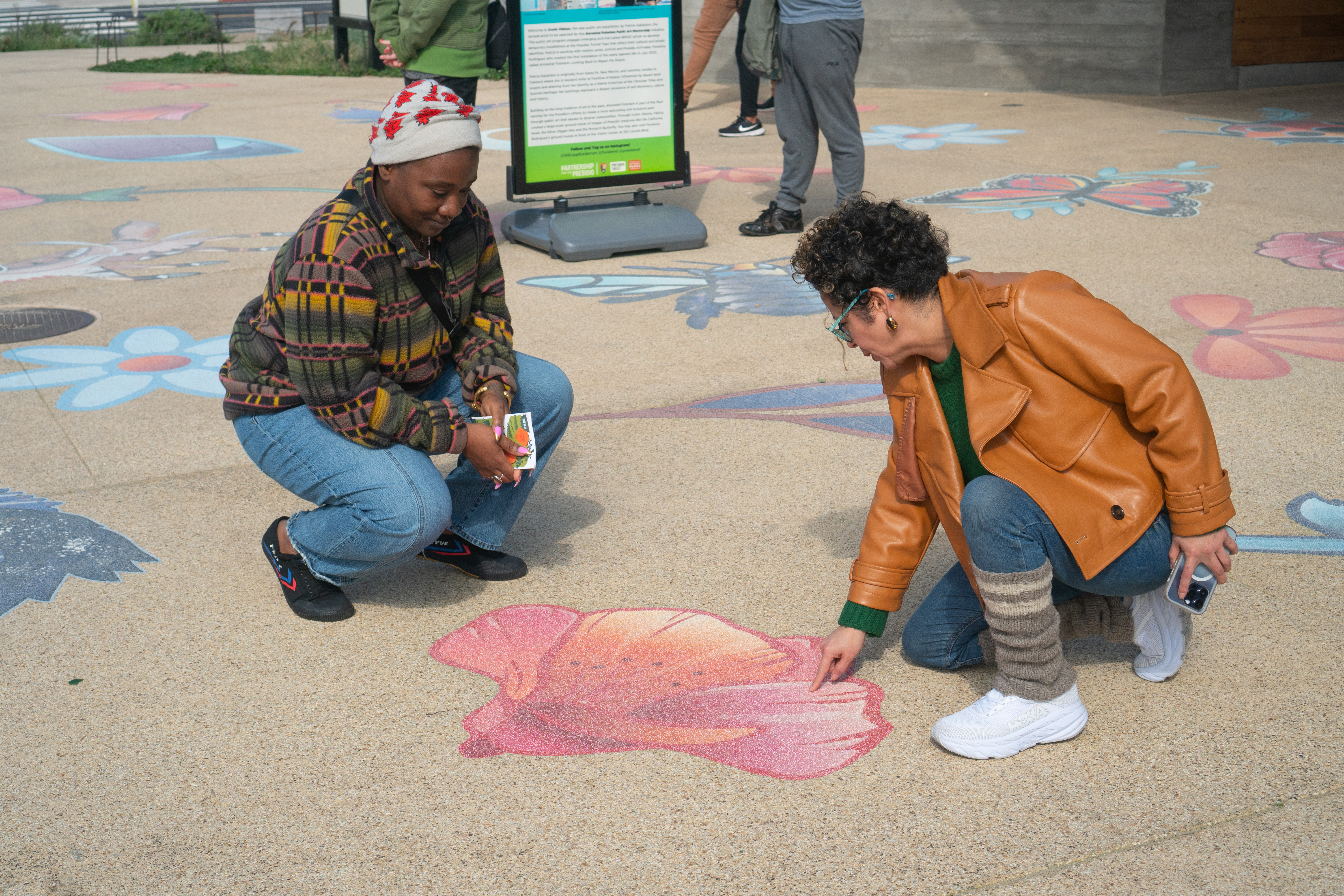 Two women, Tosha and Artist and Presidio Activator Favianna Rodriguez, kneeling over artwork on the cement at Presidio Tunnel Tops.
