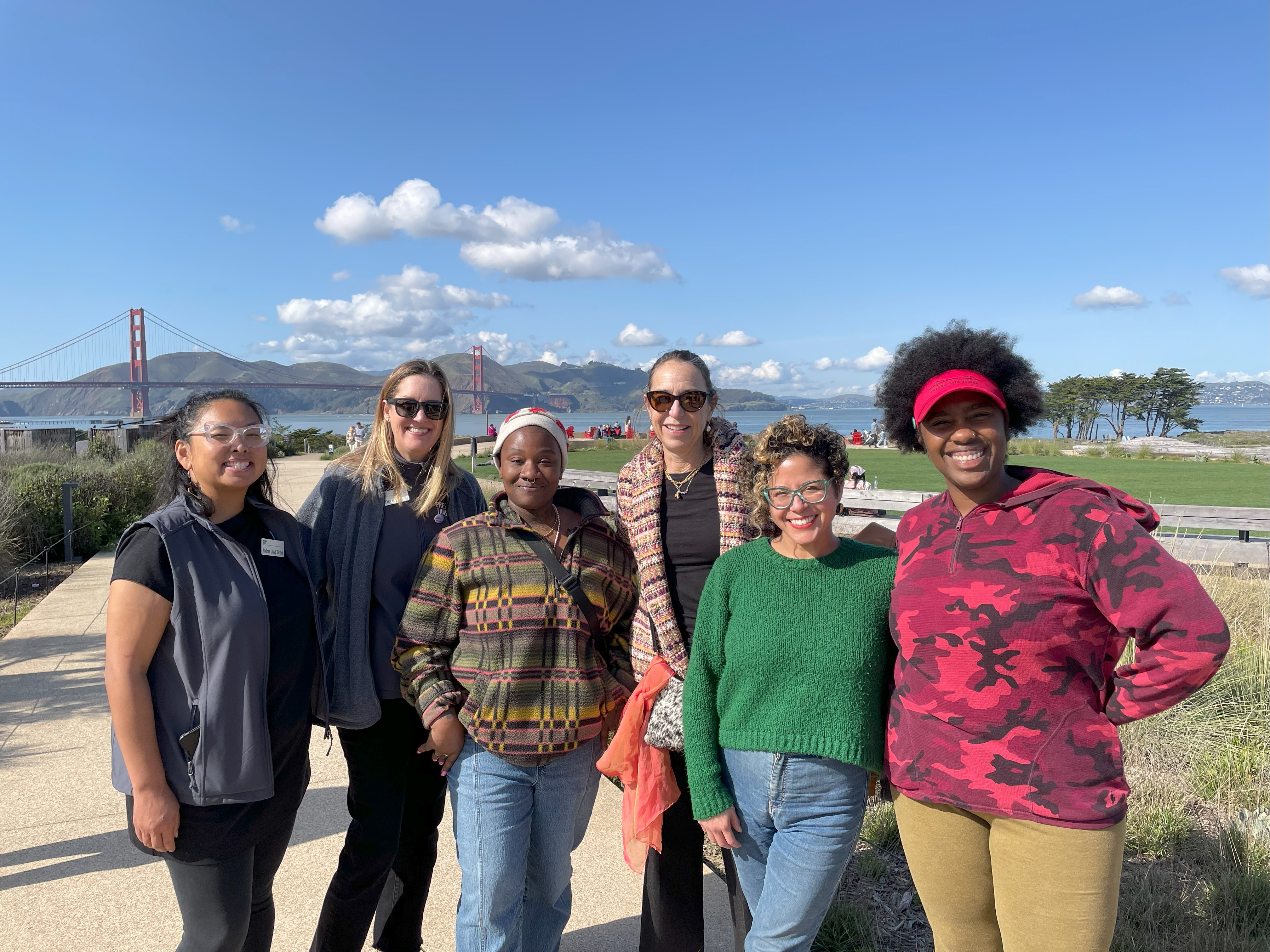 A group of women, Tosha with Favianna and representatives from the Presidio’s Community Partnership team, standing and smiling on the Presidio Tunnel Tops with the Golden Gate Bridge in the background.