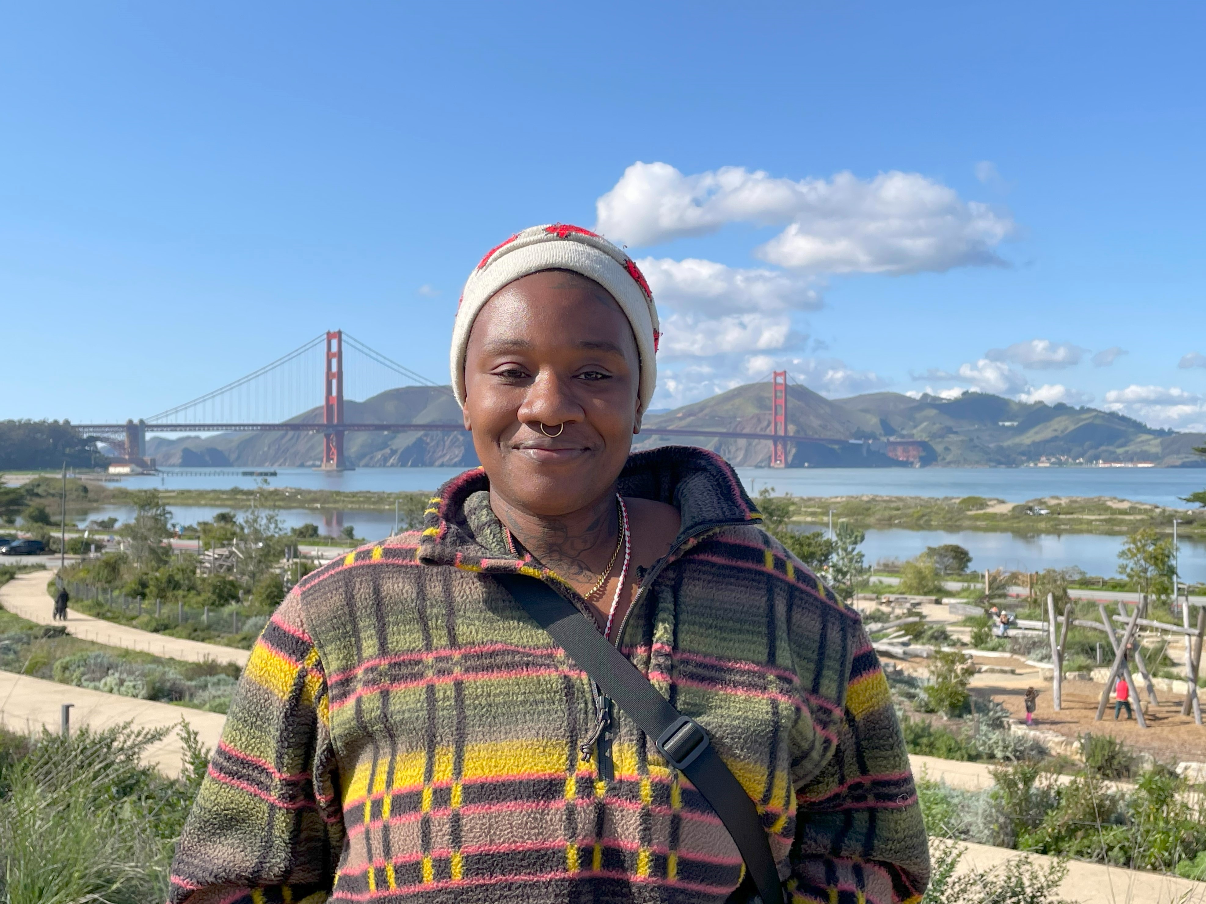 Image of a woman standing and smiling in a plaid jacket on the Presidio Tunnel Tops with the Golden Gate Bridge in the background.