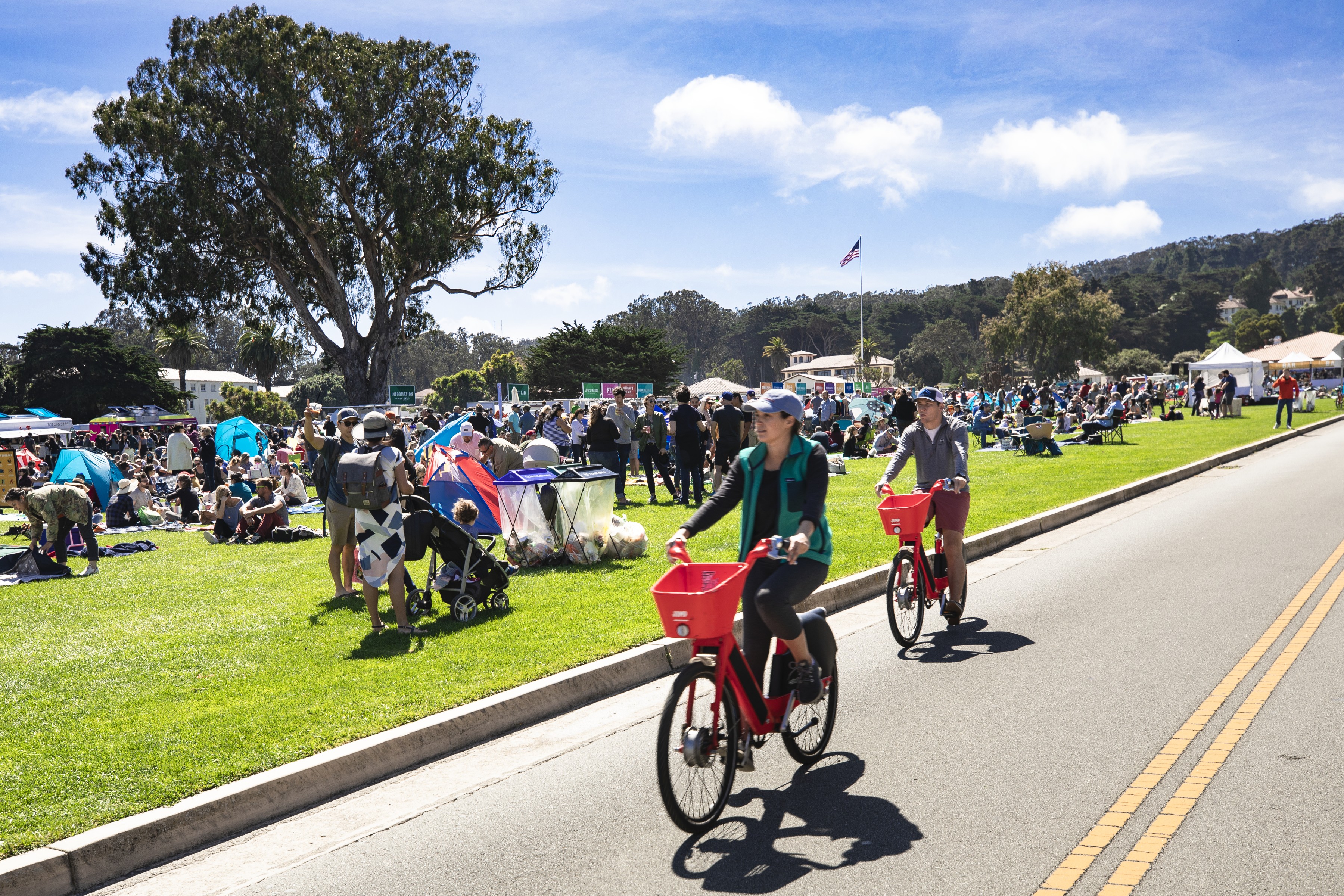 Couple biking along main parade lawn with crowd of people enjoying picnics behind them